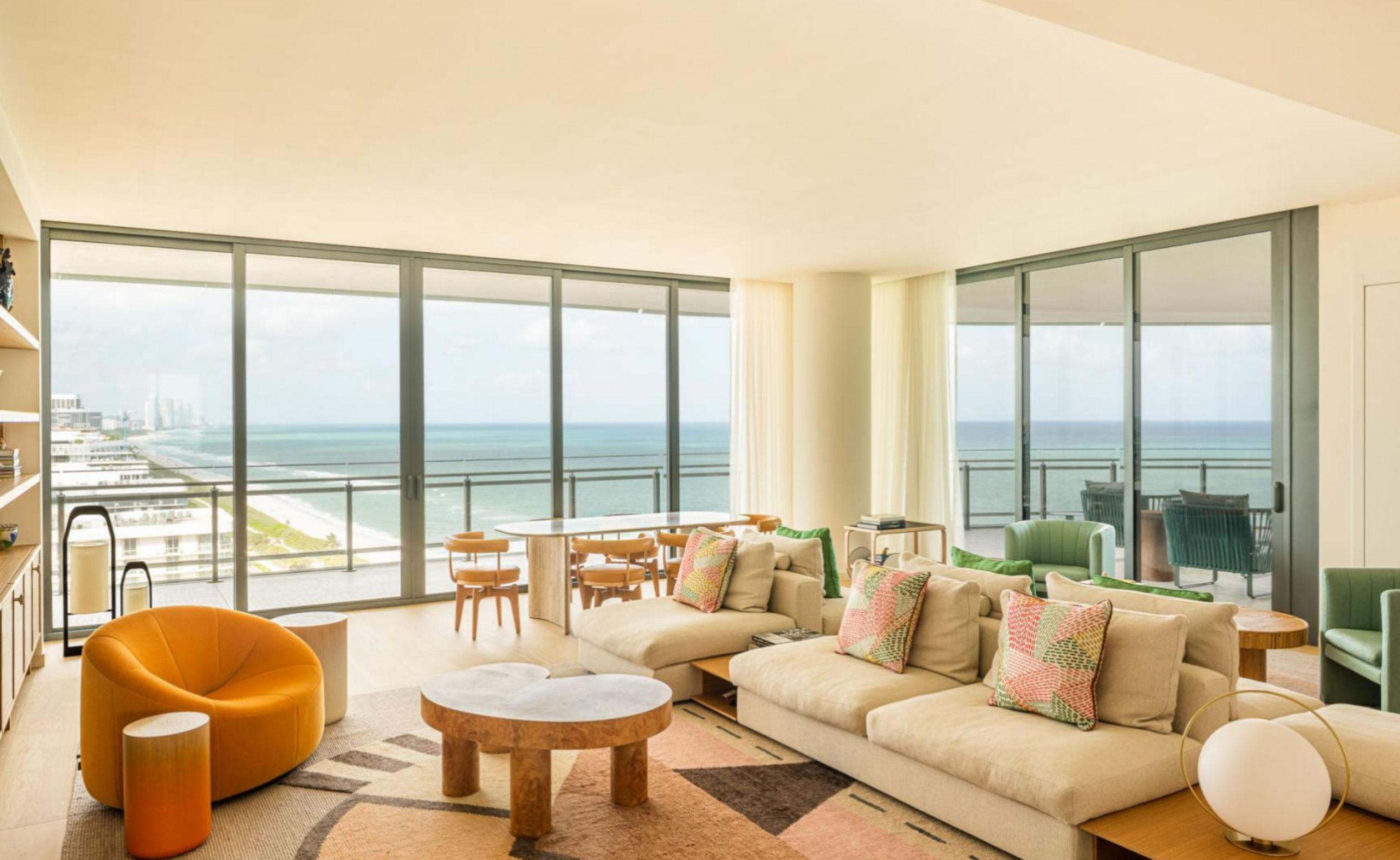 Bright living room with floor-to-ceiling windows, ocean views, neutral-toned sofas with colorful pillows, modern accent chairs, and a sculptural coffee table. Build out by Troy Dean Home.