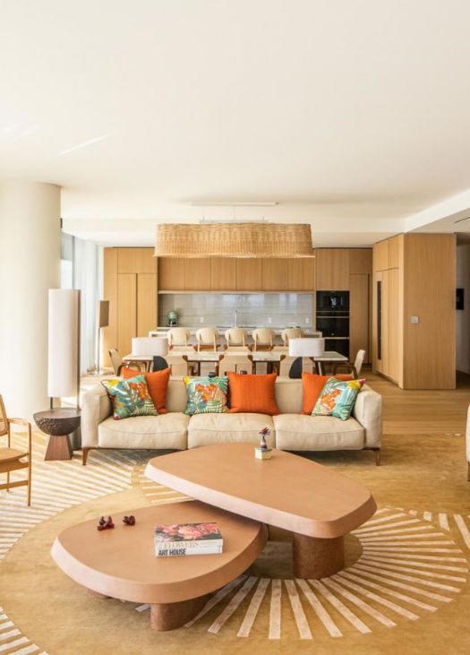 Open-concept living and dining area with neutral tones, colorful accent pillows, modern wooden furniture, and a sleek kitchen in the background. Build out by Troy Dean Home.